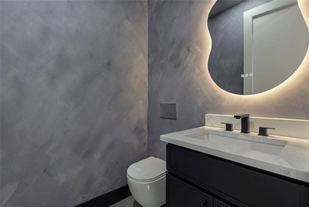 Textured grey wall finish, floating vanity with stone countertop, matte black widespread faucet, back-lit organic-shaped mirror, and wall-mounted toilet