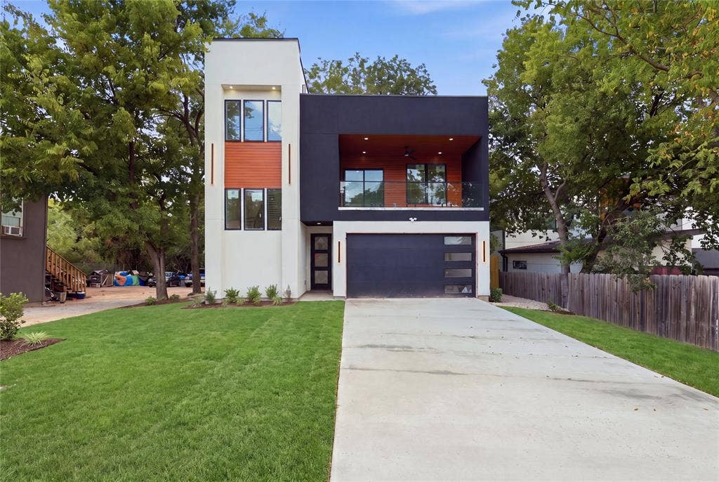 Modern two-story exterior featuring high-contrast stucco siding, wood-toned accents, a glass-railed second-level balcony, and a multi-panel garage door