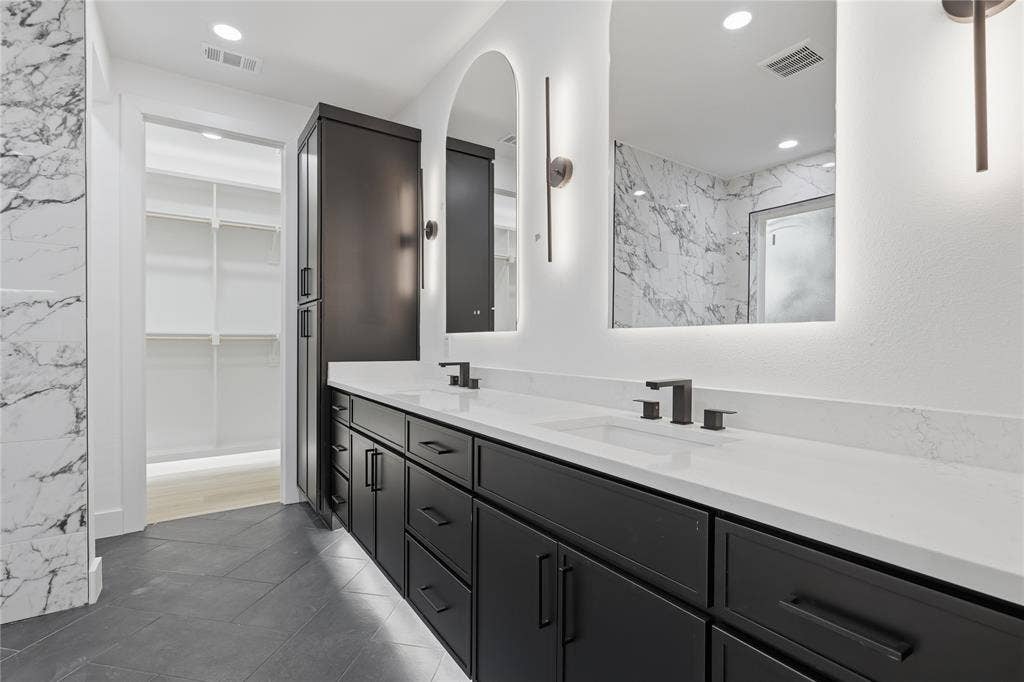 Dual vanity featuring dark cabinetry, quartz countertops, undermount sinks, matte black fixtures, and arched mirrors with backlighting, complemented by large-format tile flooring and stone-finish wall cladding