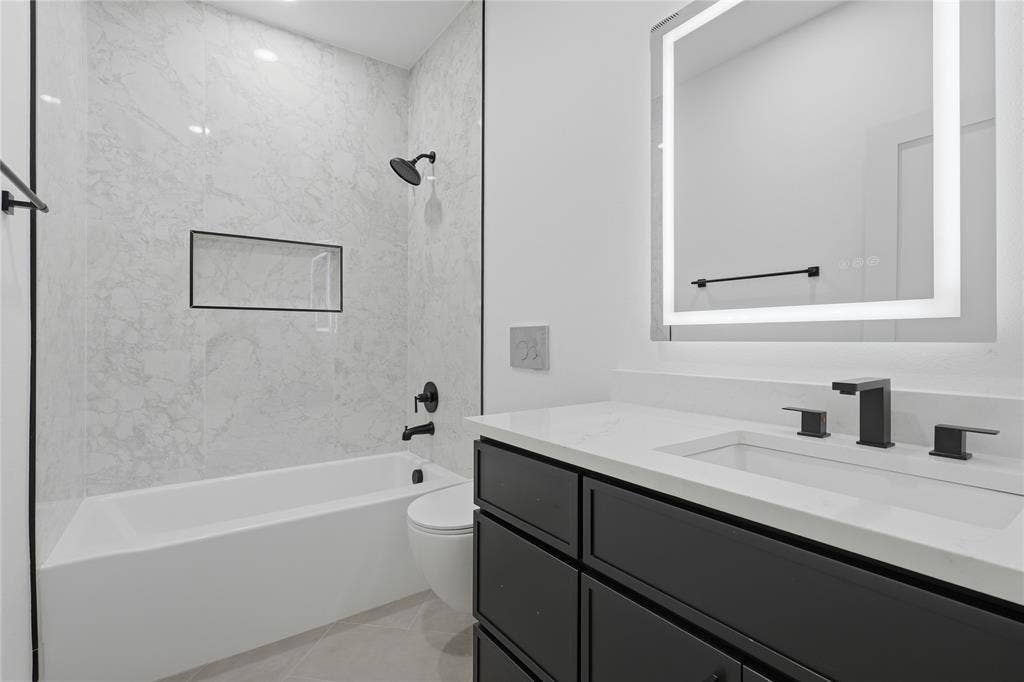 Bathroom featuring a vanity with a white countertop and dark cabinetry, an illuminated mirror, and a soaking tub with stone-patterned wall tile, a recessed niche, and matte black fixtures