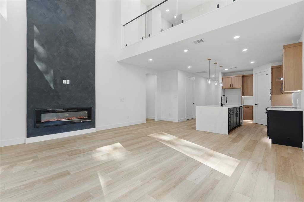 Double-height living area featuring a floor-to-ceiling dark textured fireplace surround with a linear electric insert, wood-finish flooring, recessed lighting, and a glass-paneled mezzanine railing