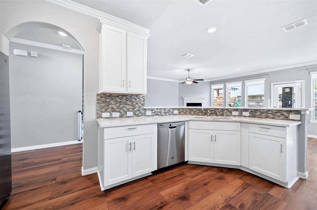 Kitchen featuring white shaker-style cabinetry, a multi-tone tile backsplash, stainless steel dishwasher, stone-finish countertops, and wood-finish flooring