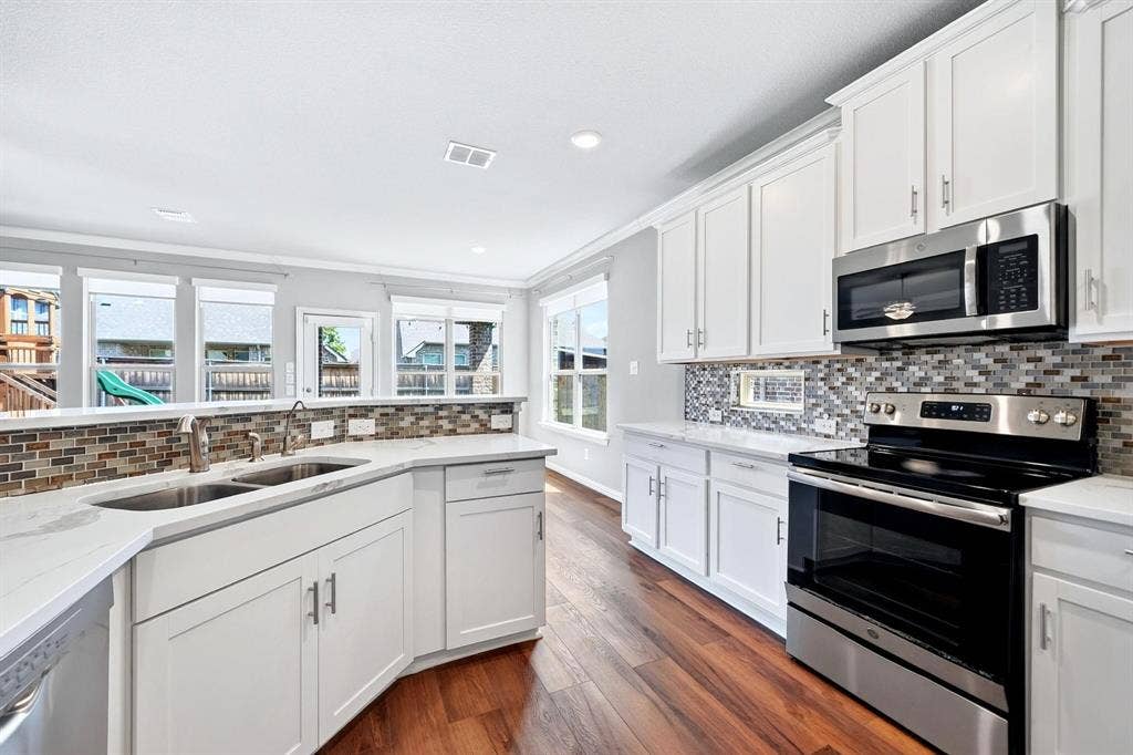 Kitchen featuring white shaker-style cabinetry, stainless steel appliances, multi-tonal glass tile backsplash, light-colored countertops, and wood-finish flooring