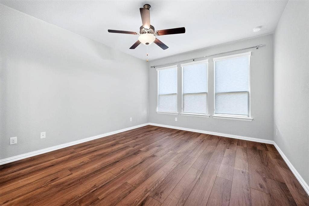Room featuring wood-finish flooring, three windows with roller shades, and a ceiling-mounted fan with integrated lighting