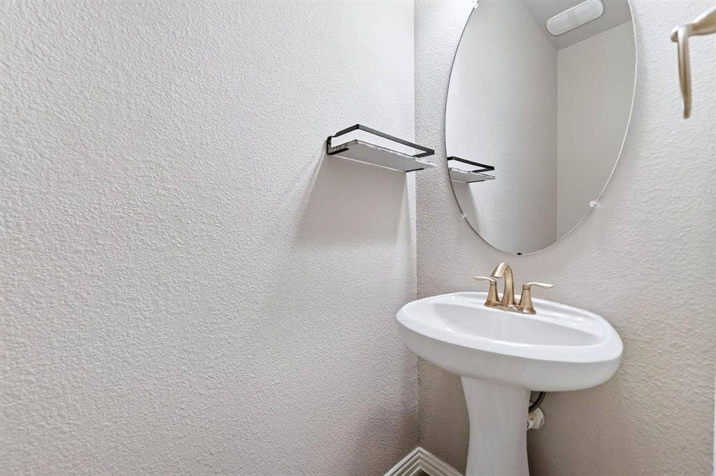 White pedestal sink with a metallic finish dual-handle faucet, oval frameless wall mirror, and a wall-mounted metal accessory shelf