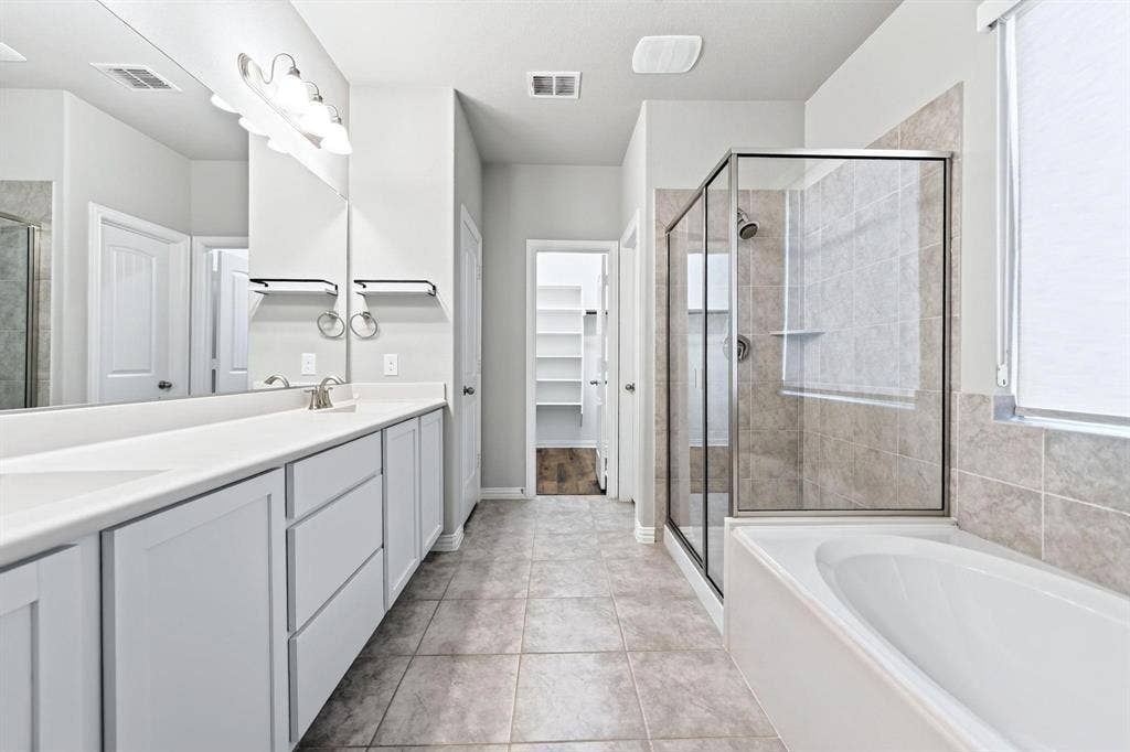 Bathroom featuring a dual-sink vanity with white cabinetry, a glass-enclosed shower, a soaking tub, and tile flooring
