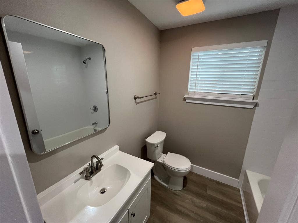 1104 Wake Drive, Richardson, TX 75081 Bathroom featuring wood-finish flooring, a white vanity with an integrated sink and metal faucet, a large rectangular mirror with rounded corners, and a bathtub with a shower fixture