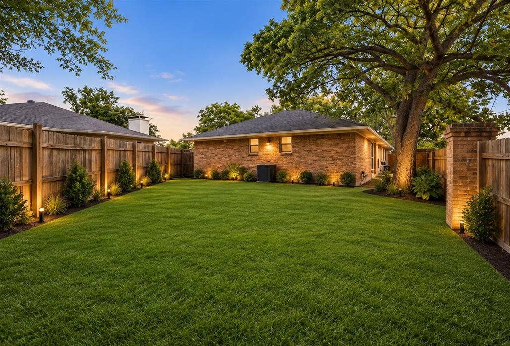 Expansive backyard featuring a brick exterior, mature oak tree, perimeter wood fencing, and installed low-voltage landscape lighting