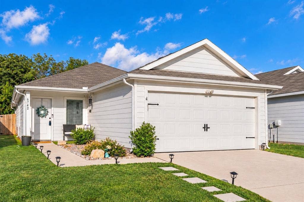 6742 Highlands Street, Princeton, TX 75407 Single-story exterior featuring white horizontal siding, a gabled roofline, an attached two-car garage with decorative hardware, and a paved concrete driveway