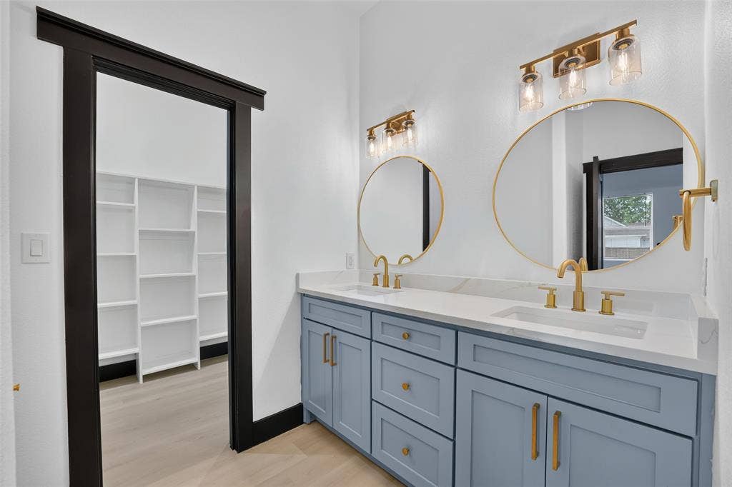 601 Melody Circle, Kaufman, TX 75142 Dual-sink vanity with light blue cabinetry, gold-tone hardware, quartz countertops, round gold-framed mirrors, and brass-finish wall lighting