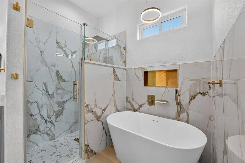 601 Melody Circle, Kaufman, TX 75142 Freestanding soaking tub with gold-tone wall-mounted hardware, glass-enclosed shower with large-format stone-patterned wall tile, recessed tiled shower niche, and circular LED pendant lighting