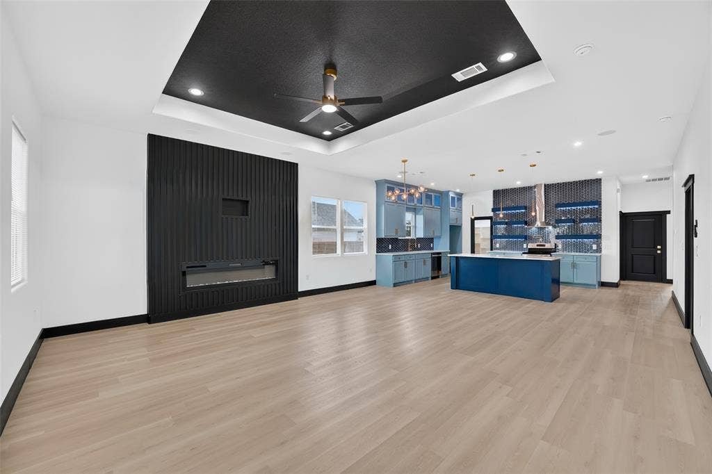 601 Melody Circle, Kaufman, TX 75142 Open-concept interior featuring light wood-finish flooring, a tray ceiling with a black accent inset, a floor-to-ceiling vertical-slat fireplace surround, a central kitchen island with blue cabinetry, and a geometric-tiled backsplash