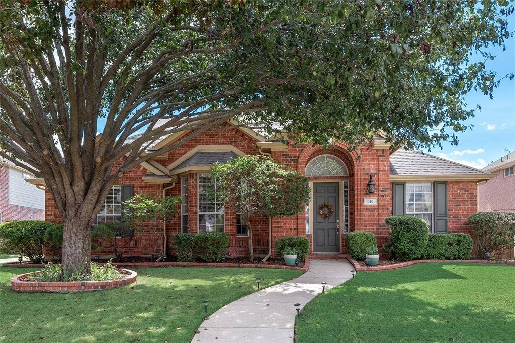 148 Oakbluff Drive, Murphy, TX 75094 Brick exterior featuring an arched entryway, a bay window, a manicured lawn, and a mature tree with a circular brick planter