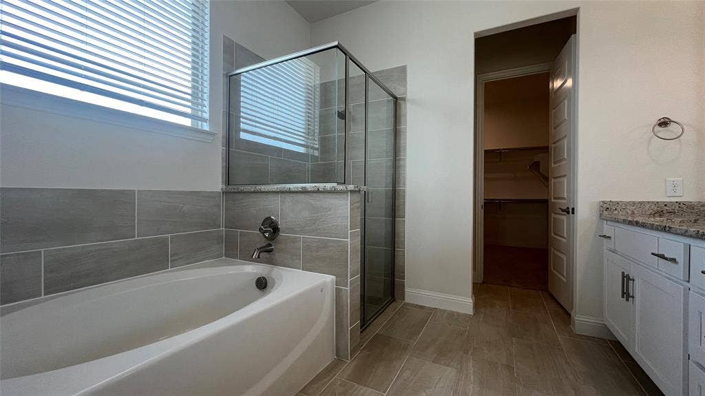 333 Elk Drive, Fate, TX 75189 Soaking tub with horizontal tile surround, glass-enclosed shower, stone-finish vanity countertop, white shaker-style cabinetry, and tile flooring
