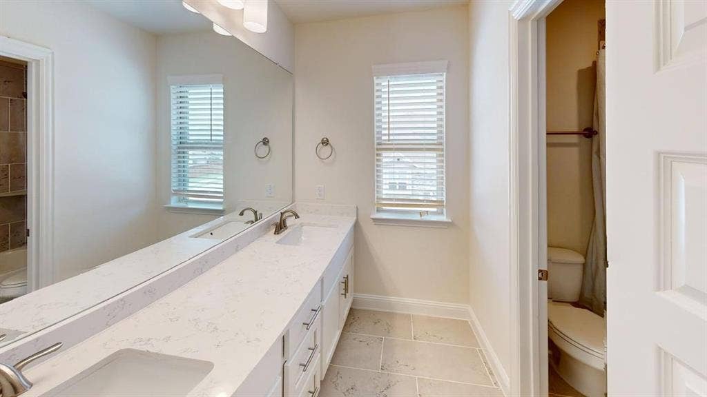 333 Elk Drive, Fate, TX 75189 Dual-sink vanity with white stone-finish countertop, full-width wall mirror, tile flooring, and dedicated enclosed water closet