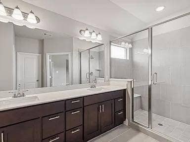 333 Elk Drive, Fate, TX 75189 Dark-finish cabinetry featuring dual undermount sinks, stone countertops, expansive wall-to-wall mirror, glass-enclosed shower with built-in bench, and tile flooring