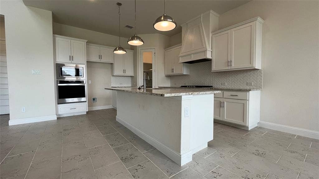 333 Elk Drive, Fate, TX 75189 Kitchen featuring white cabinetry, stone countertops, a center island with a built-in sink, stainless steel wall-mounted oven and microwave, and herringbone tile backsplash