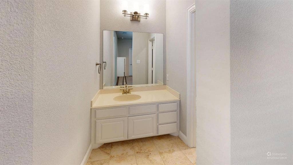 7005 Golf Club Drive, McKinney, TX 75070 Bathroom vanity featuring a solid surface countertop, an integrated oval basin, light-toned cabinetry, a wall-mounted mirror, and stone-tile flooring