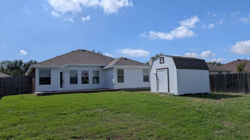 408 Oxford Loop Loop, Princeton, TX 75407 Rear exterior featuring a white siding finish, asphalt shingle roof, expansive lawn, detached storage structure, and wood privacy fencing