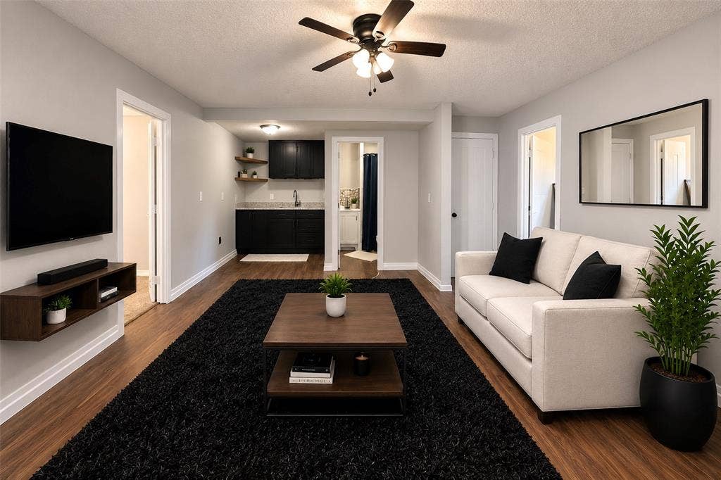 Living area featuring wood-finish flooring, a ceiling fan with integrated lighting, and a kitchenette equipped with black cabinetry, stone-style countertops, and open wood shelving