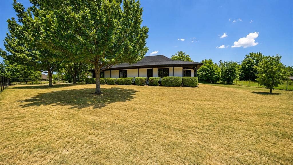 Single-story residence featuring a light-colored exterior, dark-toned roof, wrap-around covered porch, and mature shade trees situated on a sprawling lot