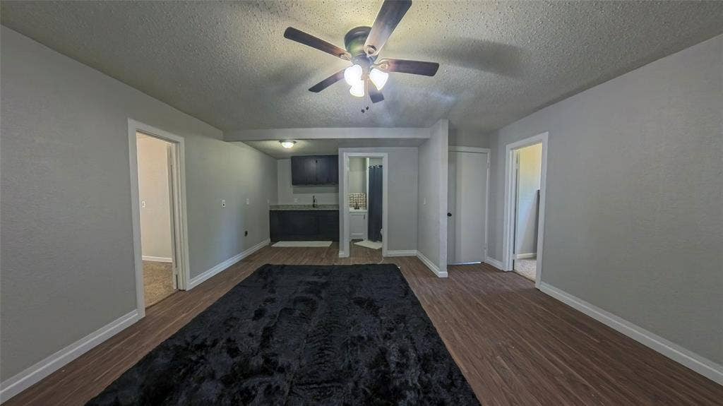 Room featuring wood-finish flooring, a ceiling fan with integrated lighting, a kitchenette with dark cabinetry, and neutral wall tones