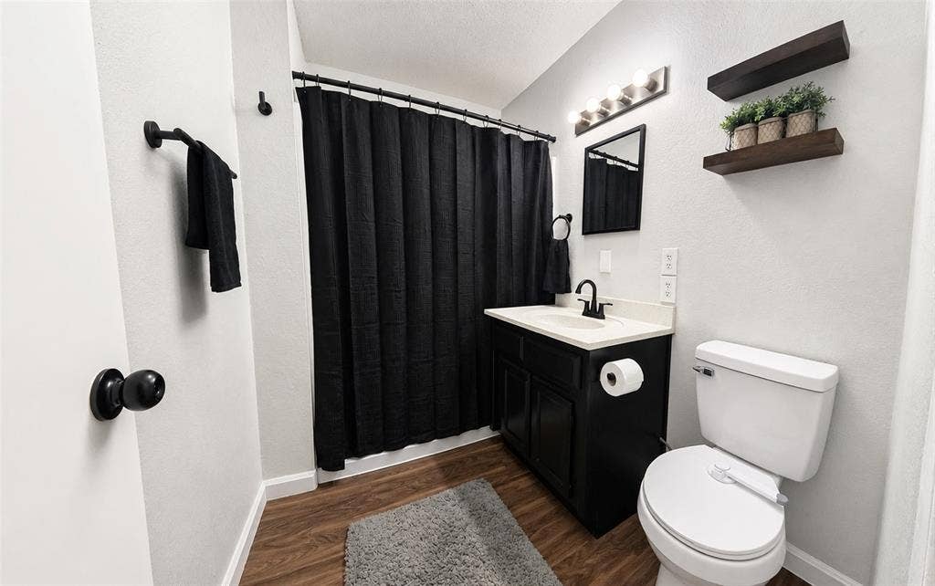 Bathroom featuring wood-finish flooring, a black vanity cabinet with a stone-top integrated sink, matte black faucet hardware, and wall-mounted floating wood shelving