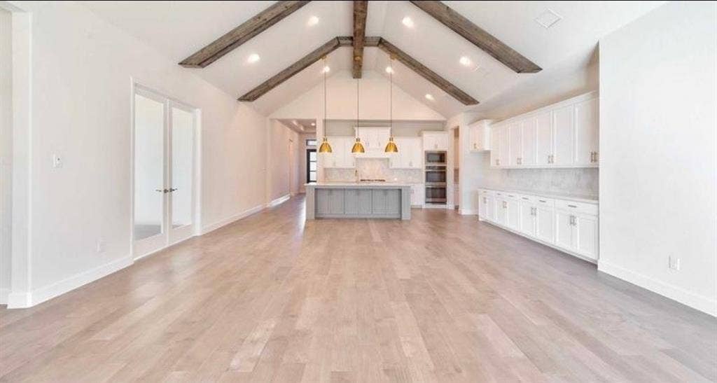 Vaulted ceiling with exposed wood beams, wood-finish flooring, central kitchen island with pendant lighting, and extensive custom cabinetry