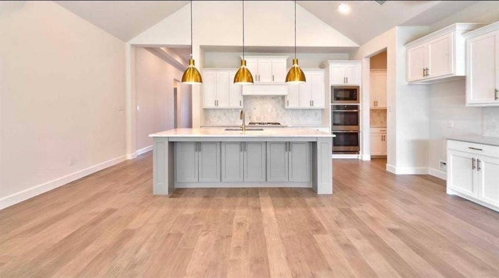 Vaulted ceilings, kitchen island with stone countertops, wood-finish flooring, shaker-style cabinetry, and recessed wall-mounted ovens