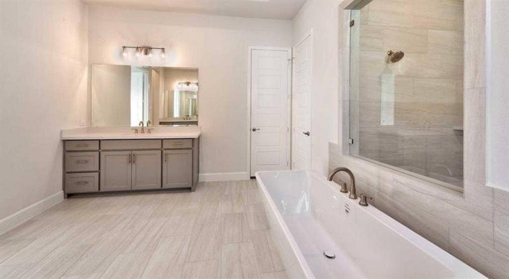 Freestanding soaking tub with a deck-mounted faucet, walk-in tile shower with a glass enclosure, dual-sink vanity with light-toned cabinetry, and large-format tile flooring