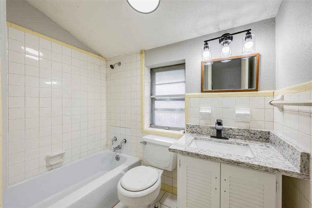 Bathroom featuring a vanity with a stone-finish countertop, white louvered cabinetry, wall-mounted three-light fixture, and tile-surround tub