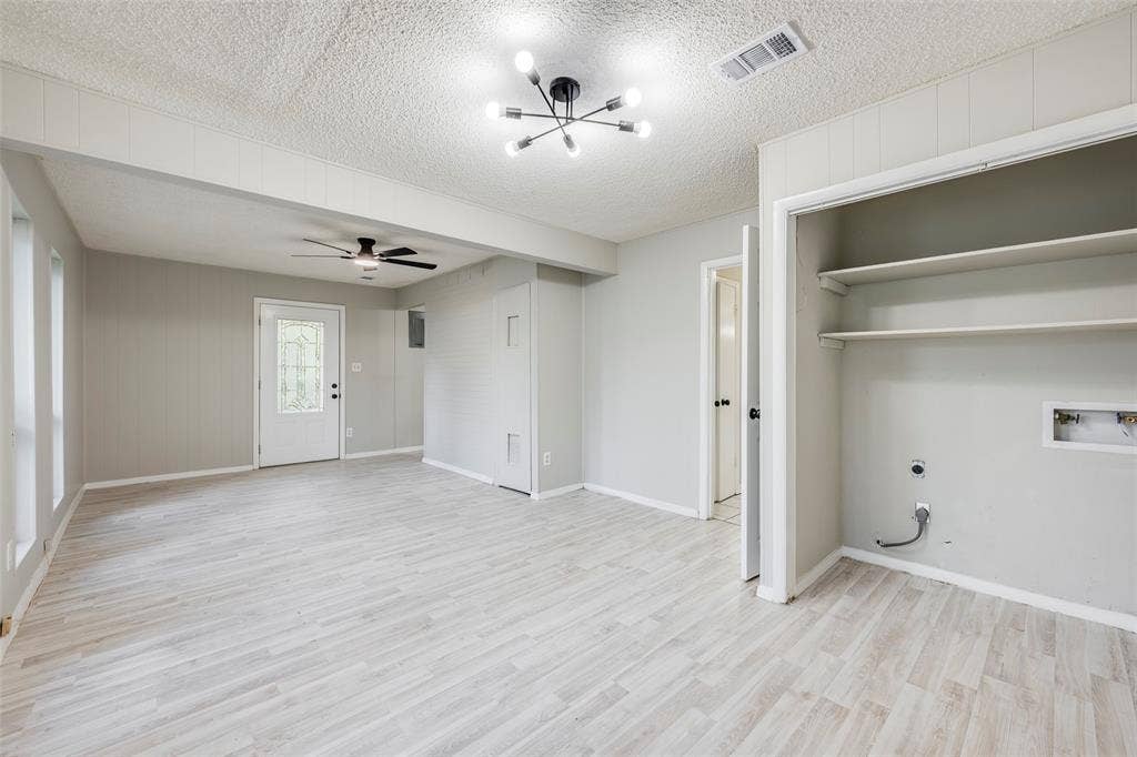 Open-concept interior featuring light wood-finish flooring, a multi-arm modern ceiling fixture, built-in wall shelving, and an exterior door with decorative glass paneling