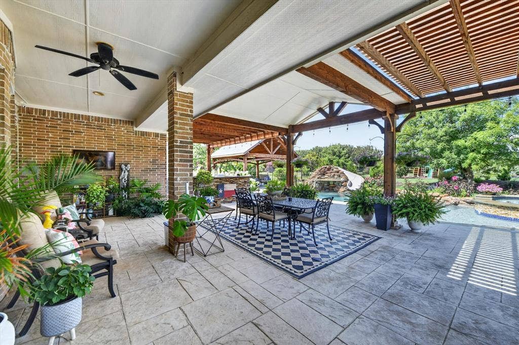Covered patio featuring stone-tile flooring, a wood-slat pergola, brick exterior walls, a ceiling fan, and access to an outdoor pool area with a water feature
