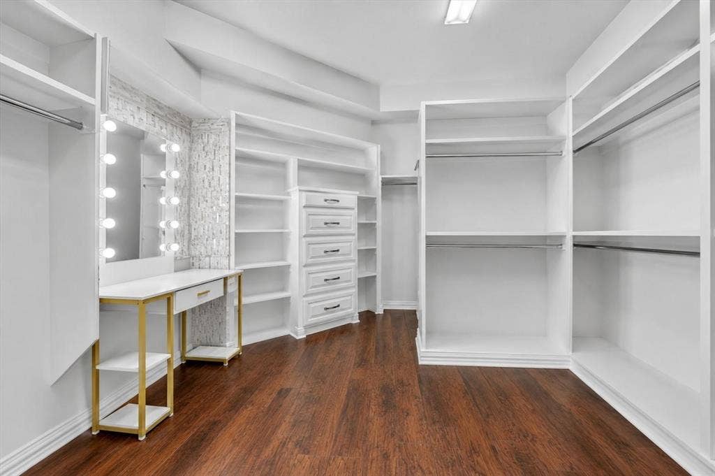 Walk-in closet featuring floor-to-ceiling white built-in shelving, integrated vanity with illuminated mirror, stacked drawer storage, wood-finish flooring, and metal hanging rods