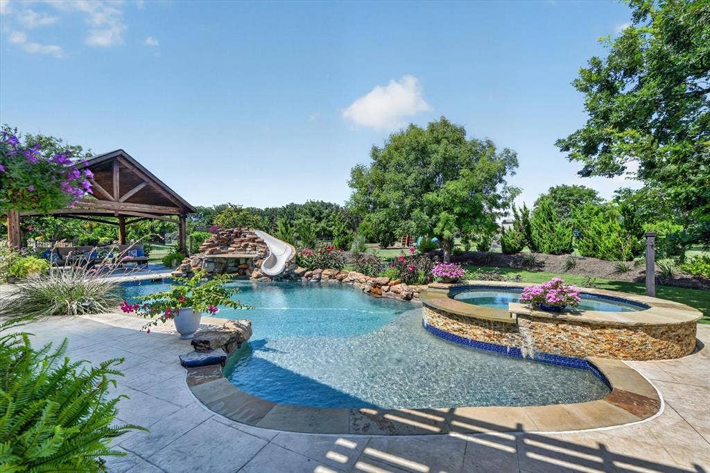 Custom swimming pool featuring a rock waterfall, integrated slide, elevated stone-clad spa, and sprawling stamped concrete patio with a detached timber-frame pavilion