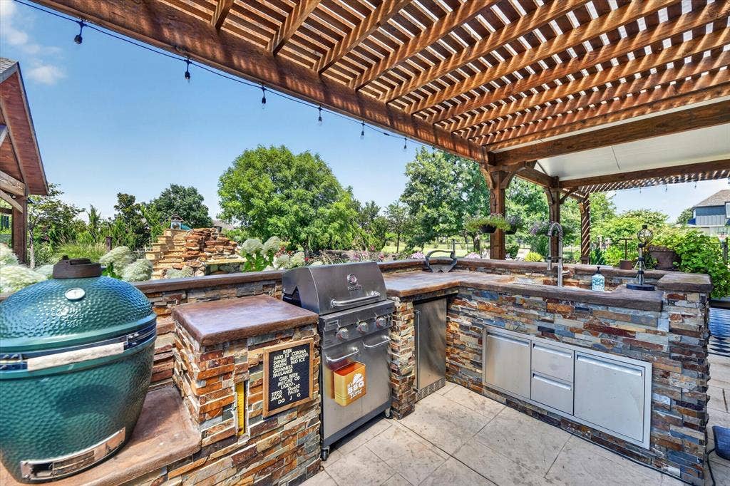 Outdoor kitchen featuring a stacked stone exterior, concrete countertops, stainless steel appliances, a built-in sink, and a cedar wood pergola