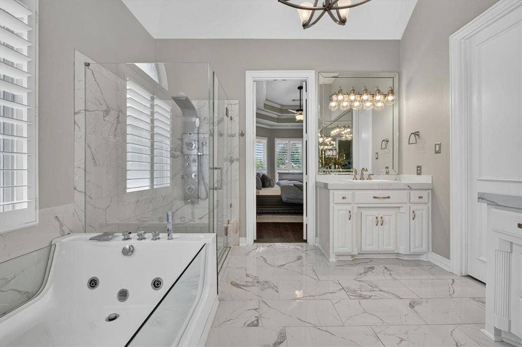 Bathroom featuring large-format tile flooring, jetted soaking tub, glass-enclosed shower with body spray system, white cabinetry with stone countertops, and a wall-to-wall vanity mirror with integrated lighting