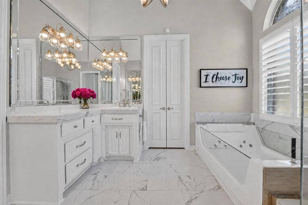 White cabinetry with light-toned countertops, stone-patterned tile flooring, full-width vanity mirror, built-in jetted tub with stone-patterned surround, and arched window with louvered shutters