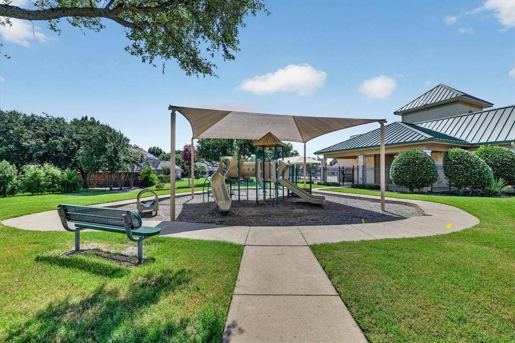 Recreational area featuring a playground structure under a large shade canopy, a concrete walkway, and a metal park bench