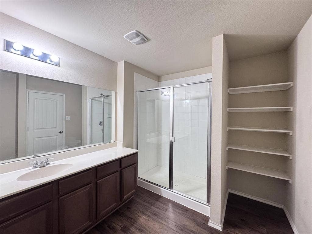 2105 Meadow Park Drive, Princeton, TX 75407 Bathroom featuring a vanity with a white countertop and oval sink, a glass-enclosed shower, wood-finish flooring, a full-width vanity mirror, and built-in wall shelving