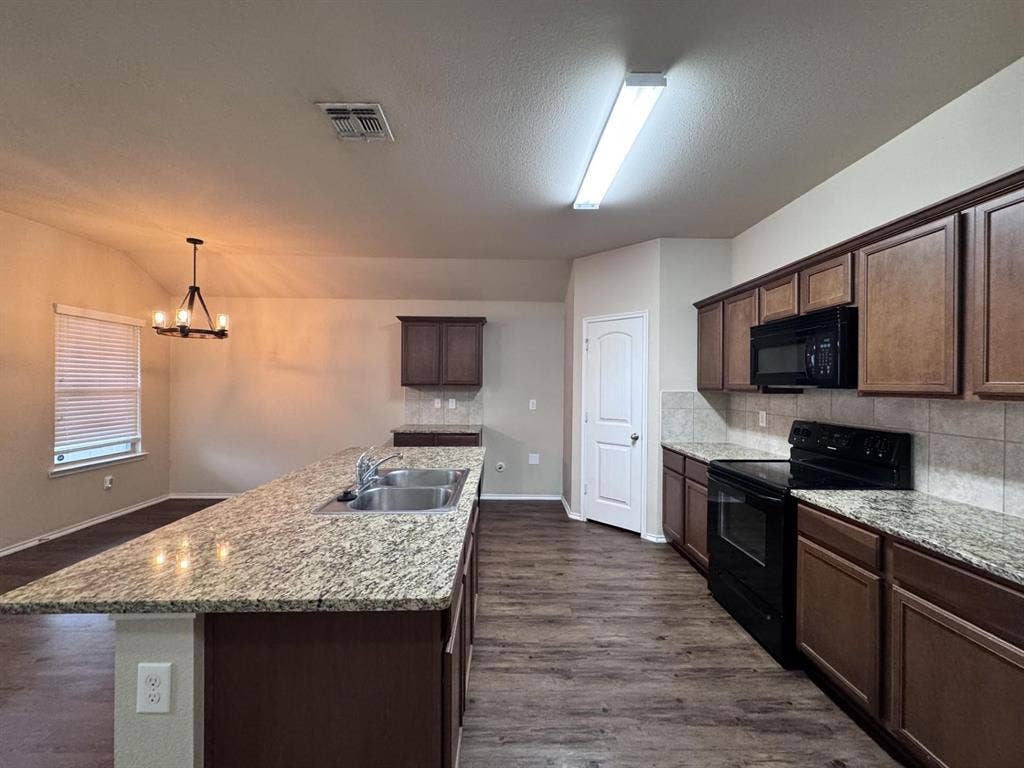 2105 Meadow Park Drive, Princeton, TX 75407 Kitchen featuring an expansive stone countertop island with a dual-basin stainless steel sink, wood cabinetry, stone-tiled backsplash, and wood-finish flooring
