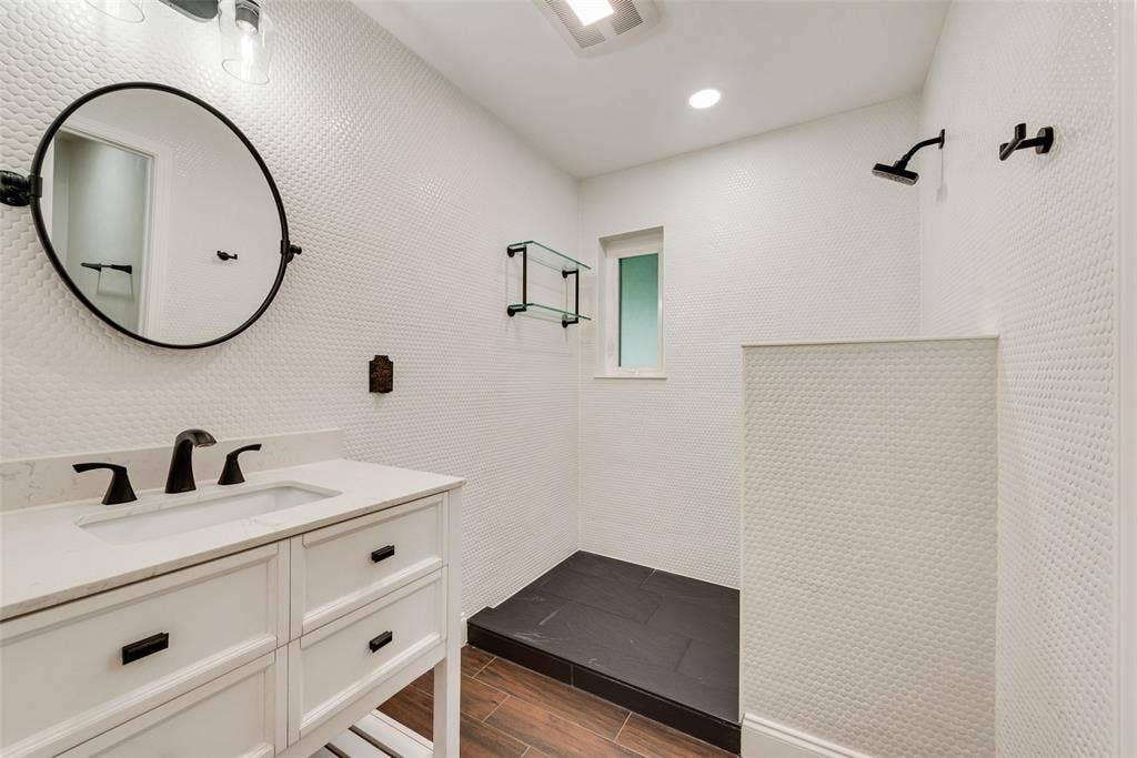 620 Cox Drive, Irving, TX 75062 White vanity featuring a stone-surface countertop, circular mirror with matte black frame, matte black plumbing fixtures, penny-tile wall cladding, and walk-in shower with dark tile flooring