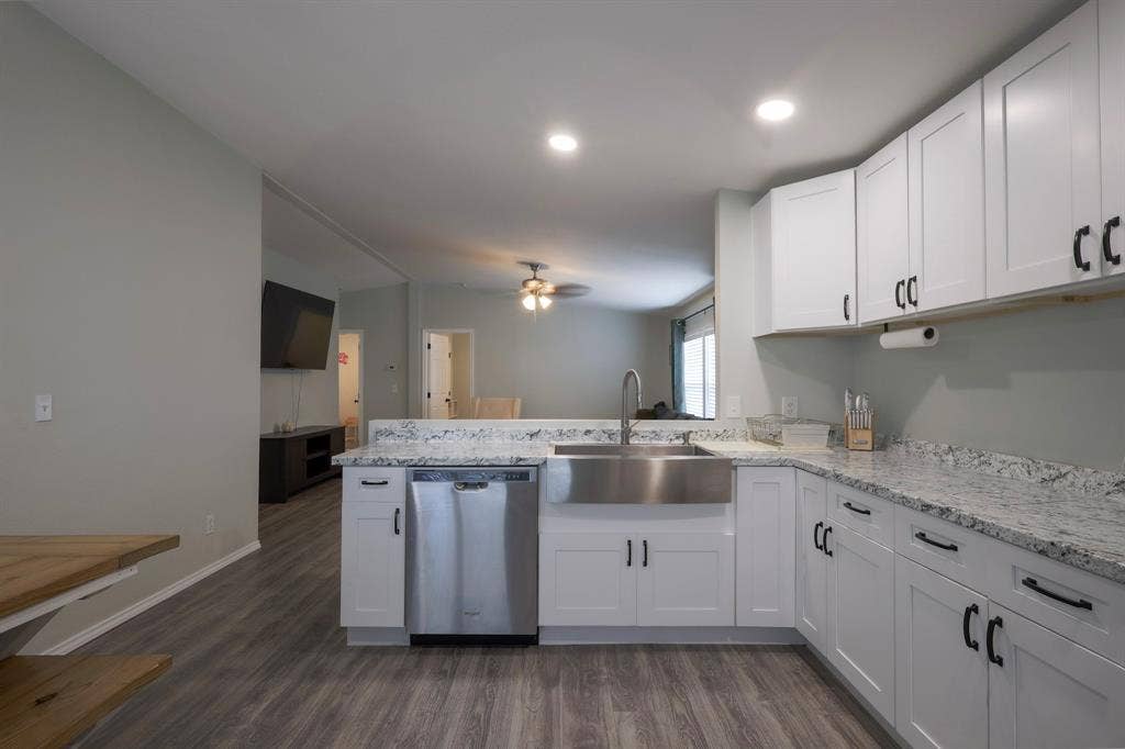Kitchen featuring white Shaker-style cabinetry, stone-finish countertops, stainless steel farmhouse sink, stainless steel dishwasher, and wood-finish flooring