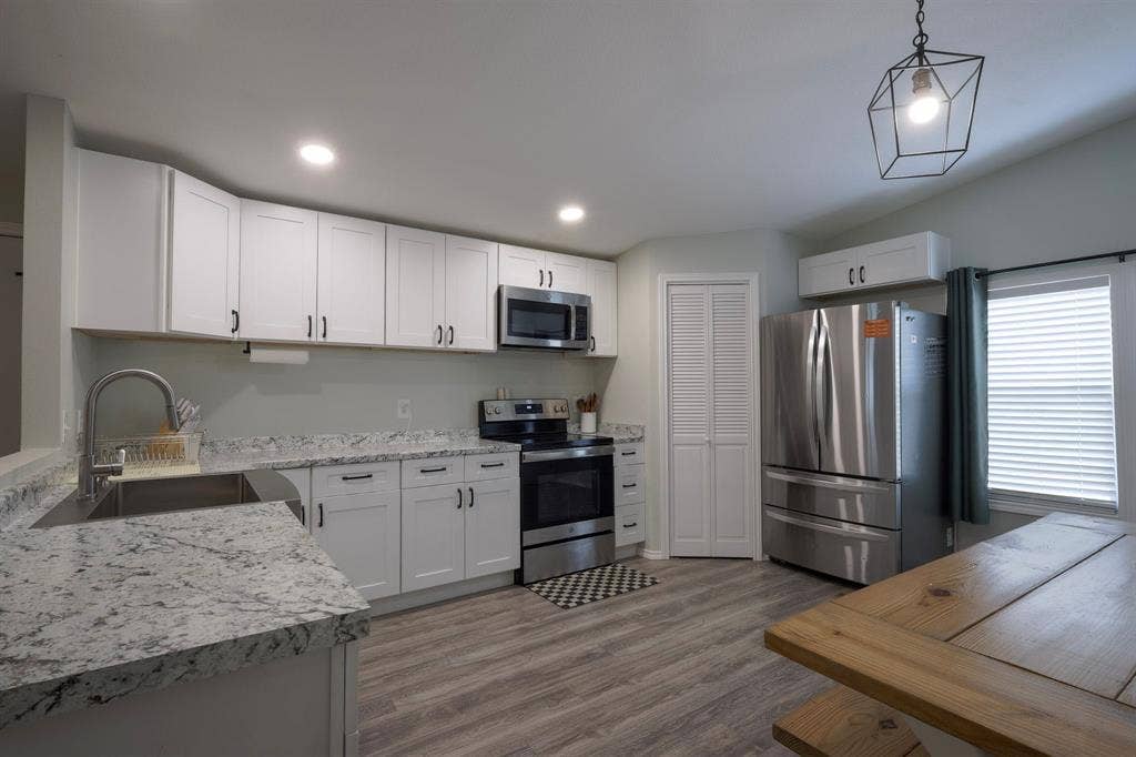 Kitchen featuring white shaker-style cabinetry, stone-patterned countertops, stainless steel appliances, wood-finish flooring, and a geometric pendant light fixture