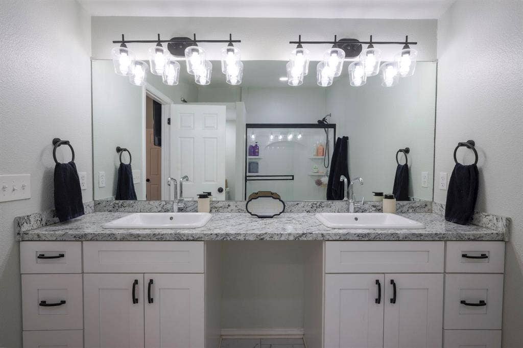 Dual-sink vanity featuring stone-finish countertops, white shaker-style cabinetry, matte black hardware, and double multi-bulb vanity lighting