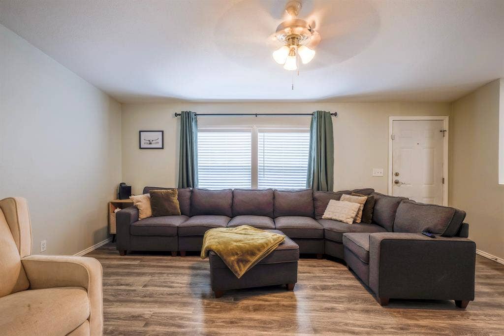 Living area featuring wood-finish flooring, a ceiling fan with multi-light fixture, and dual-pane windows with horizontal blinds