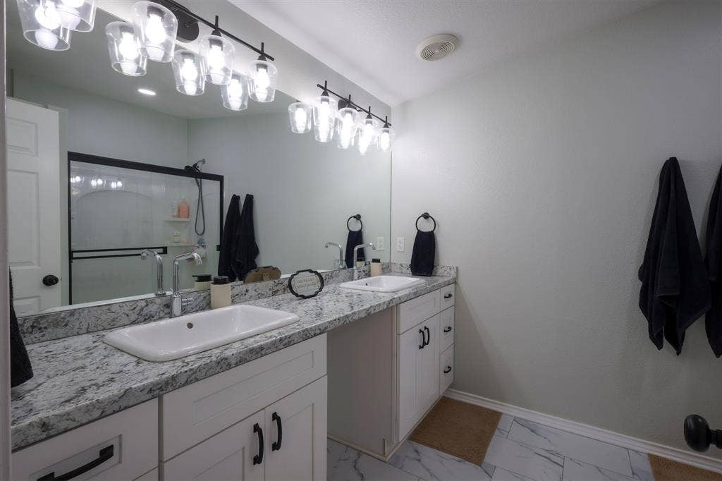 Dual-sink vanity with stone-finish countertops, undermount rectangular basins, matte black hardware, and wall-to-wall mirror with multi-bulb industrial-style lighting