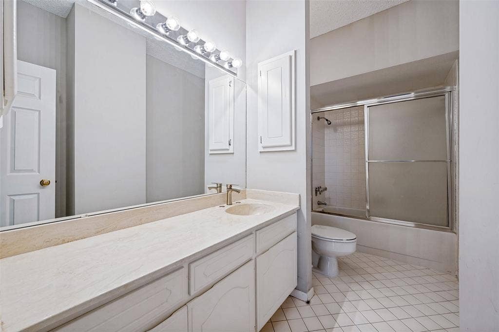 5200 Keller Springs Road, #522, Dallas, TX 75248 Bathroom vanity with a stone countertop, integrated basin, and full-width wall mirror, accompanied by a shower-tub combination featuring a sliding glass enclosure and tiled surround