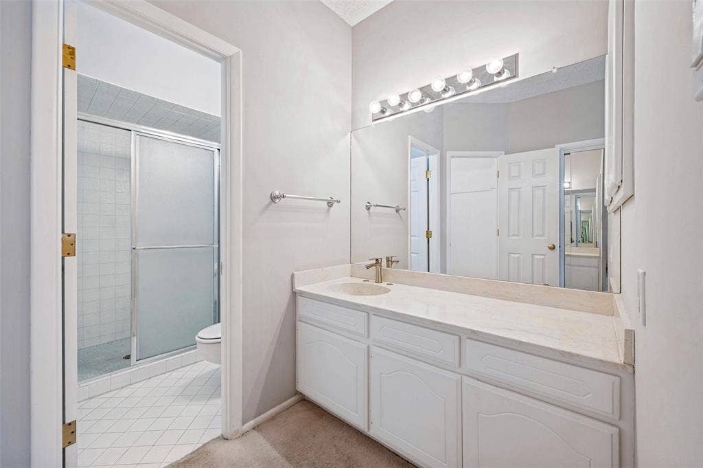 5200 Keller Springs Road, #522, Dallas, TX 75248 Bathroom featuring a white vanity with a neutral stone countertop, an expansive wall-mounted mirror, multi-bulb vanity lighting, and a glass-enclosed shower stall with tile flooring