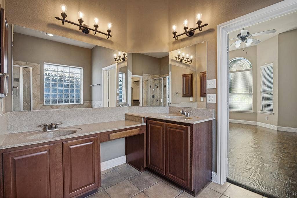 1380 Riverside Oaks Drive, Rockwall, TX 75087 Dual-vanity bathroom featuring raised-panel wood cabinetry, stone-composite countertops, multi-light vanity fixtures, and tiled flooring
